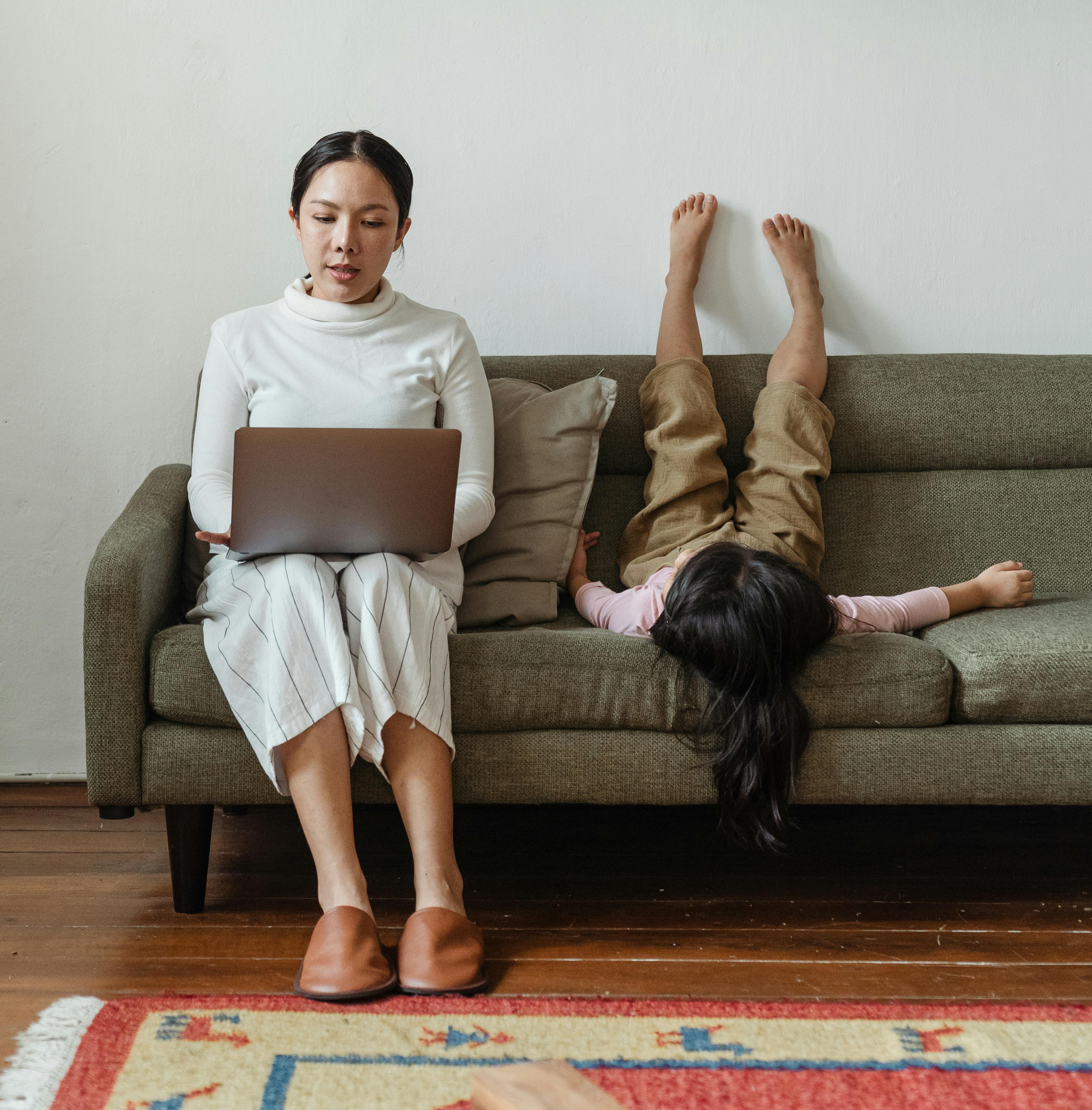 Mother typing in her laptop with her child beside her in the sofa